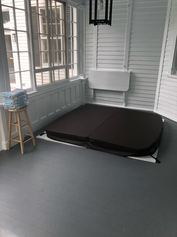 Une pièce d’angle avec un matelas sombre posé sur le sol, un tabouret en bois sur lequel sont pliés des couvertures, et une fenêtre avec des stores.