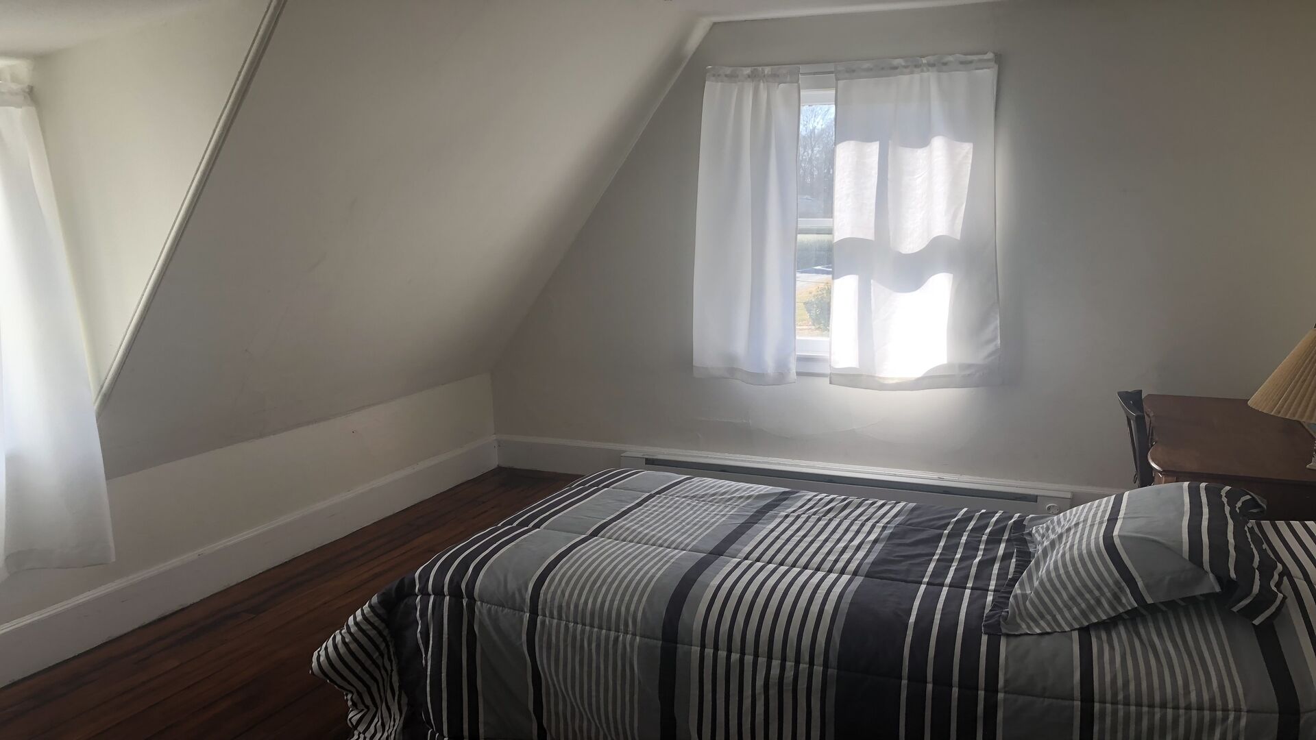 Una habitación pequeña y con techo inclinado, con una cama individual, una silla y un ventanal con cortinas.