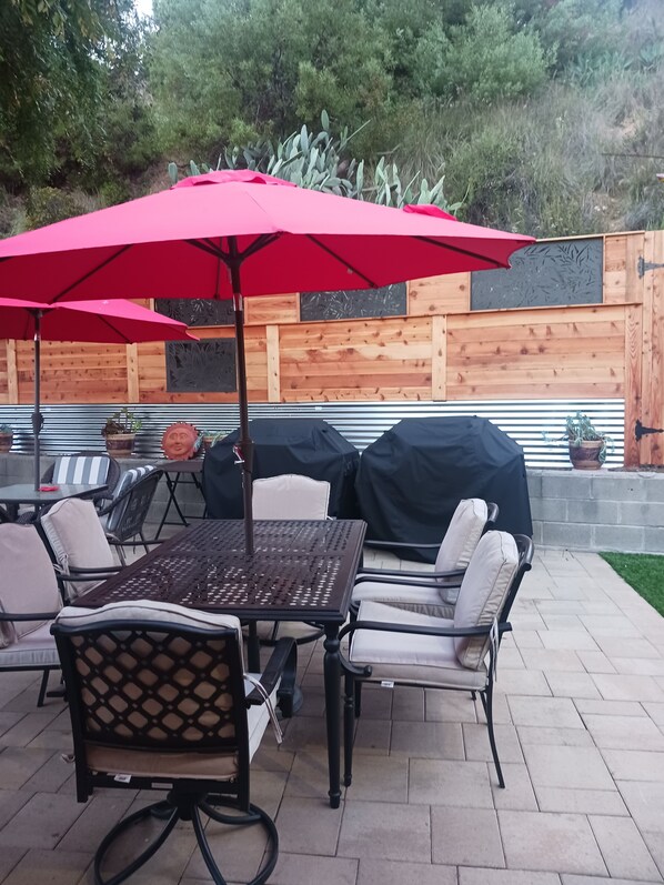 An outdoor patio with a table and chairs, a red umbrella, and a wooden fence in the background. - Private Home great single story home. Plenty of space for the family. (San Diego)
