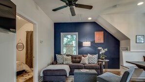 Una sala de estar moderna con una pared de acento azul oscuro, un ventilador de techo, un ventanal grande y una zona de estar acogedora con sofá y reposapiés.
