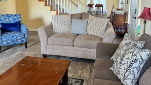 A living room with a sofa, a coffee table, and a patterned armchair. A staircase is visible in the background leading to another room.