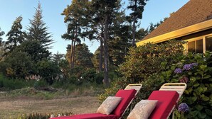 Two sun loungers with red cushions and beige pillows, positioned on a paved patio with a view of tall trees and a building with a tiled roof.
