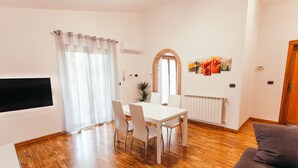 A modern living room with a wooden floor, a white dining table with four chairs, a grey sofa, and a large window with curtains. - Large appartment near the Airport, private free parking (Fiumicino)