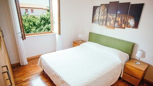 A neatly made bed with a green headboard, wooden nightstands with lamps, and a window with a view of greenery and buildings. - Large appartment near the Airport, private free parking (Fiumicino)