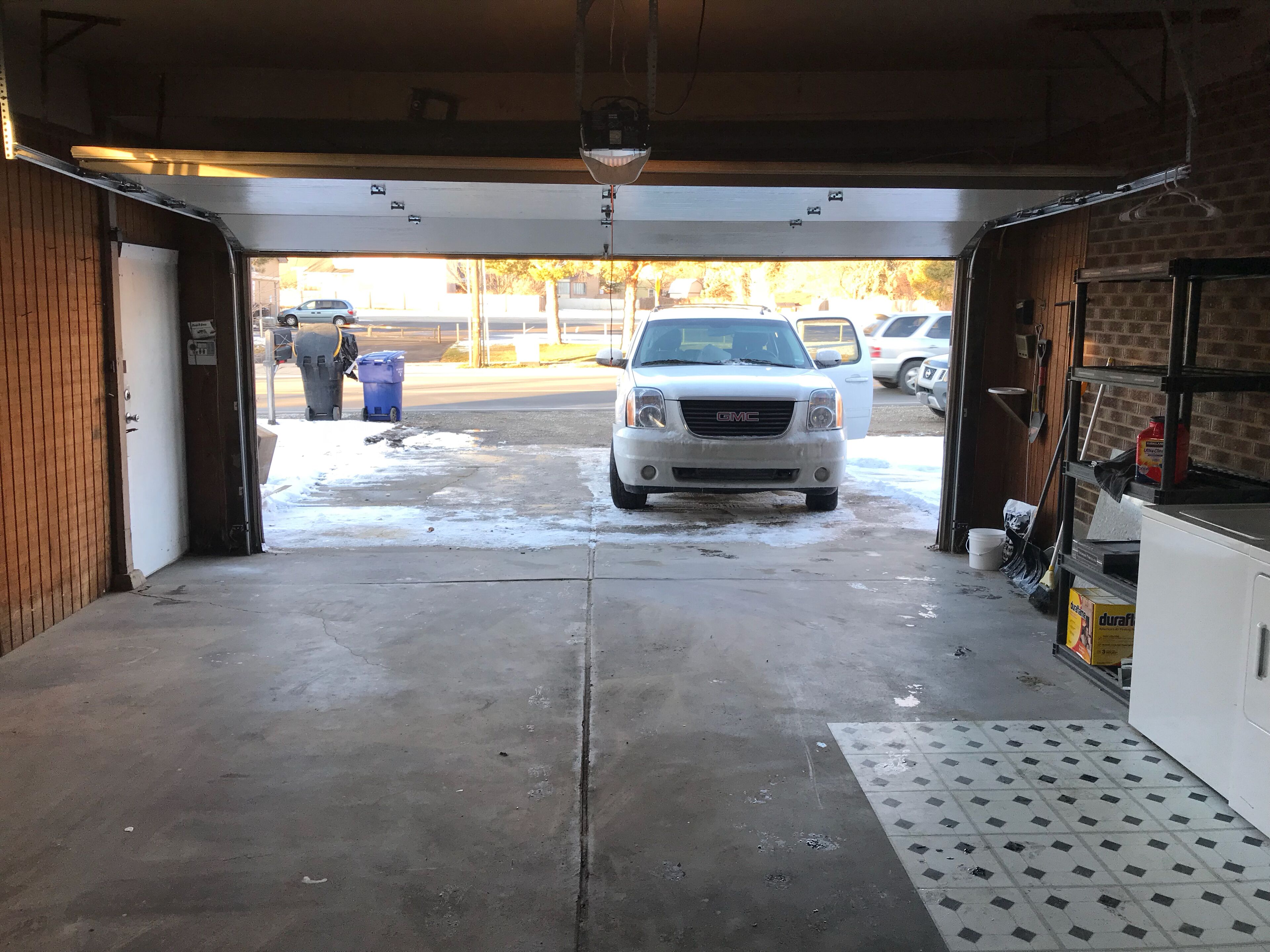 A garage with a white GMC truck parked inside, a blue trash bin, and a white utility room with shelves.
