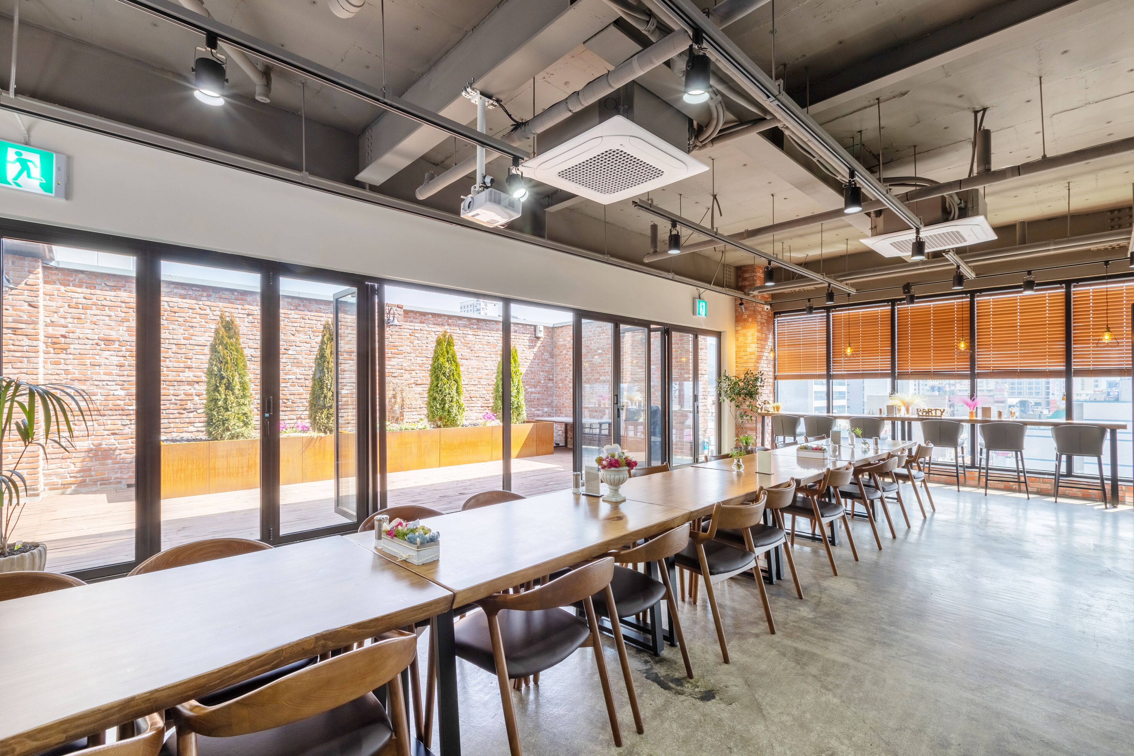 A spacious conference room with a long table, multiple chairs, and large windows offering views of a brick building and greenery.