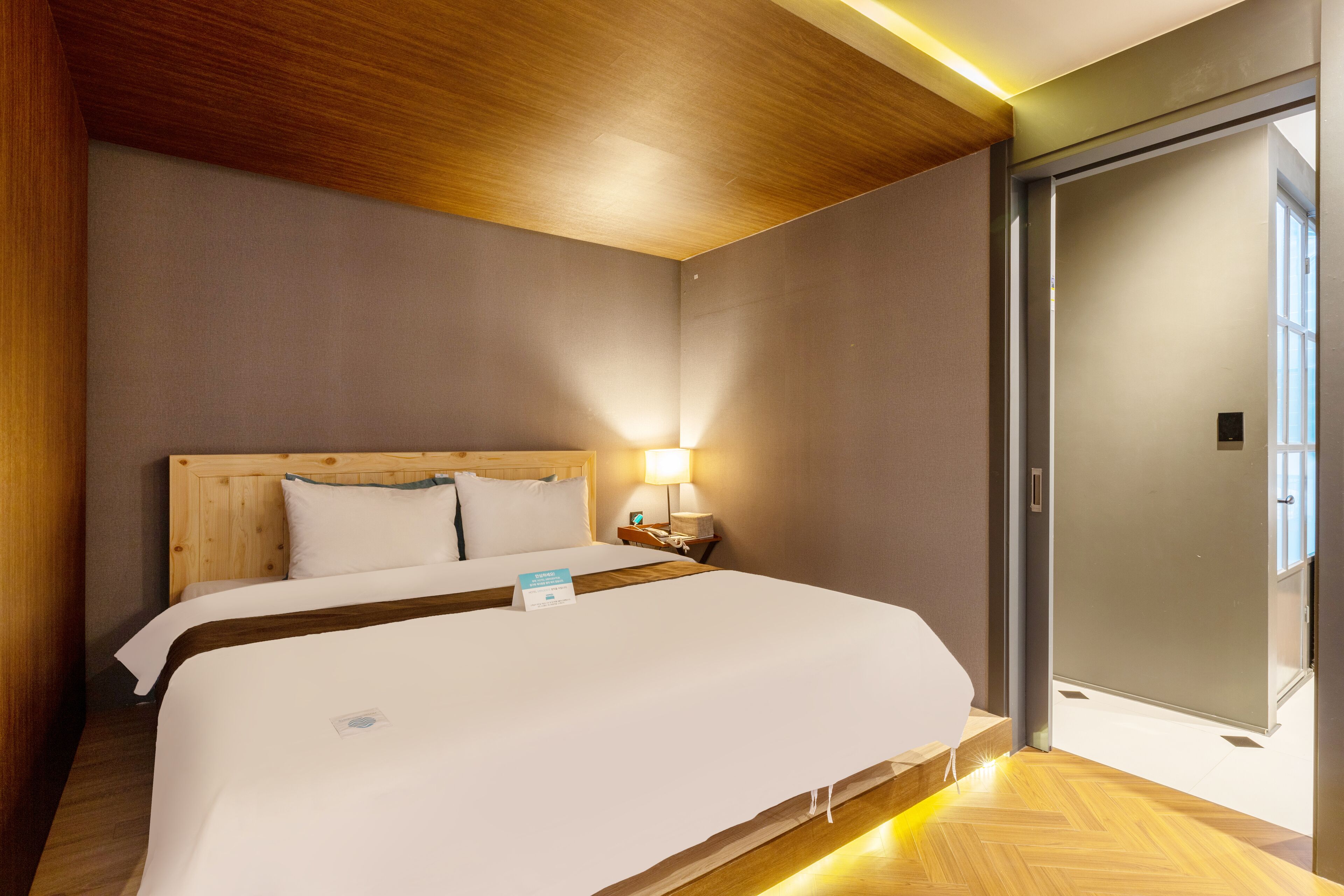 Una habitación de hotel con cabecera de madera, cama con ropa blanca, mesita de noche con lámpara y puerta que da a otra habitación.