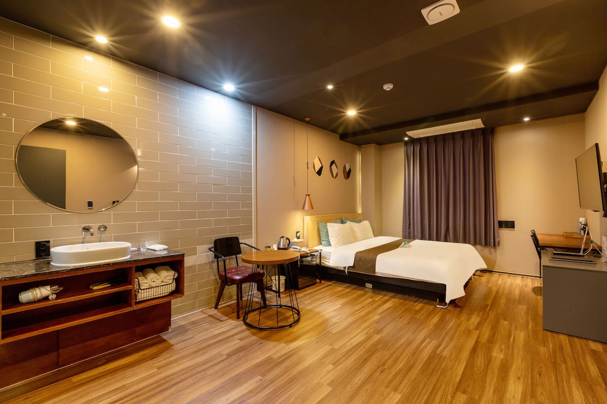 A modern hotel room with a wooden floor, a round mirror, a bed with white linens, a desk with a chair, and a small round table with a lamp.