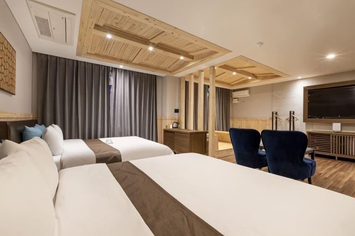 A hotel room with two beds, a desk, a chair, a TV, and a large window with curtains.