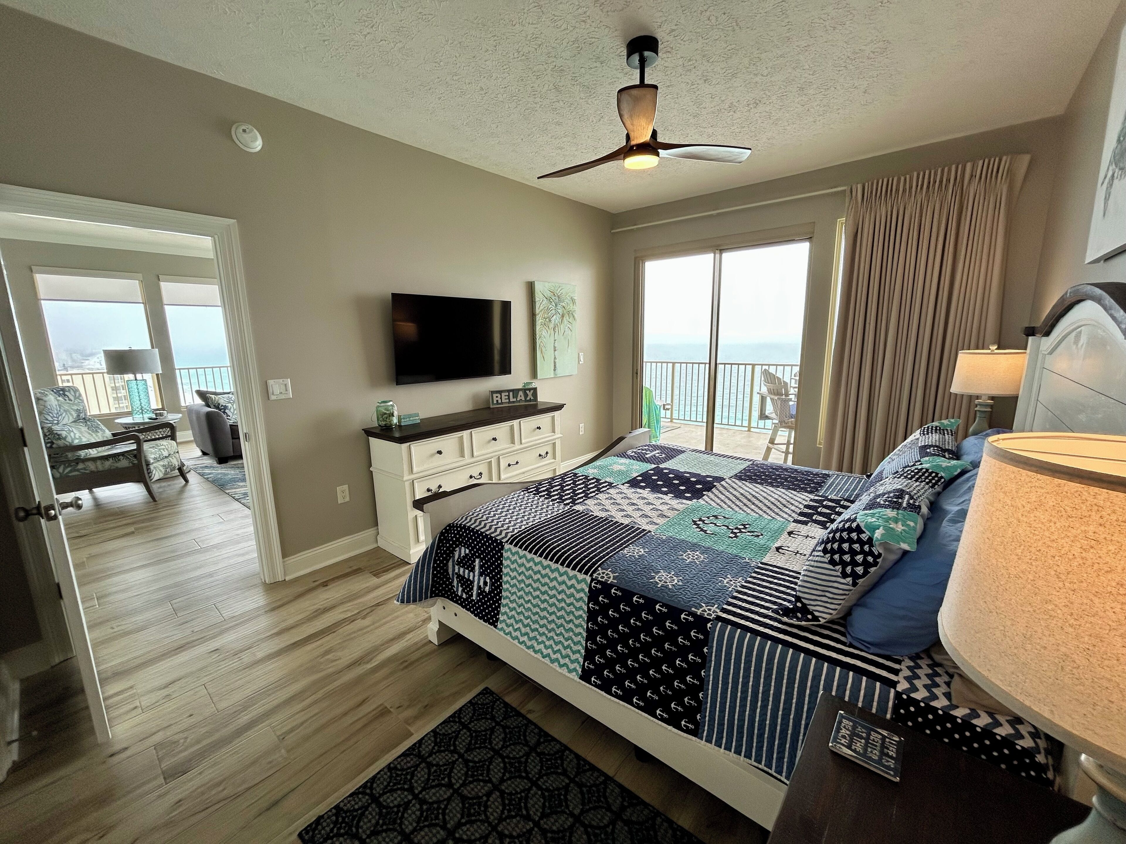A bedroom with a large bed, a ceiling fan, a TV, and a balcony with a view of the ocean.