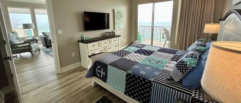 A bedroom with a large bed, a ceiling fan, a TV, and a balcony with a view of the ocean.