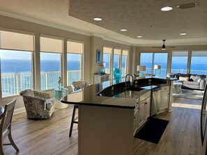 Uma cozinha ampla com uma ilha grande, sala de jantar com vista para o oceano e sala de estar com sofá e mesa de centro.