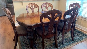 A dining area with a wooden table and chairs, a kitchen in the background, and a patterned rug on the floor.