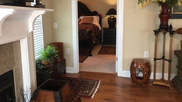 A hallway leading to a bedroom with a bed, nightstand, lamp, and a vase with yellow flowers. There is a fireplace with a plant, a wooden chair, and a small table with a vase.