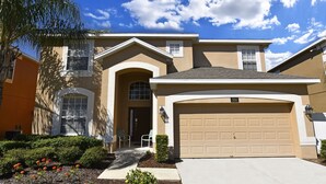 Ein zweistöckiges Haus mit einer beige farbenen Garagentür, einem Hauseingang mit einer kleinen Veranda und einem gepflegten Garten mit roten Blumen.