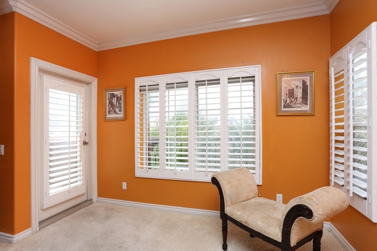 Une pièce aux murs orange, avec une porte blanche, une fenêtre dotée de volets blancs, un fauteuil beige et deux tableaux encadrés accrochés au mur.
