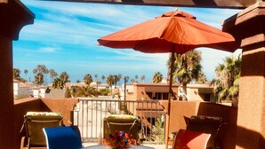 Une terrasse extérieure avec une table ronde et des chaises, un parasol rouge, et une vue sur des palmiers et des immeubles.