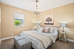 Een slaapkamer met een groot bed, twee bedlampjes, een kroonluchter, een raam met zonwering en een schilderij van een boom aan de muur.