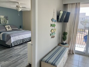Een slaapkamer met een bed, een plafondventilator, een aan de muur bevestigde televisie, een kleine potplant en uitzicht op het balkon.