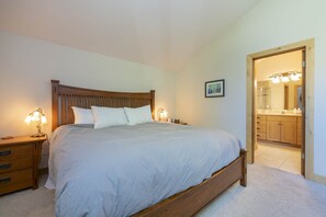 A bedroom with a wooden bed, two bedside tables with lamps, a small bedside table with a lamp, a framed picture on the wall, and a bathroom visible through an open door. - Sunrise Retreat In Sun Valley (Sun Valley)