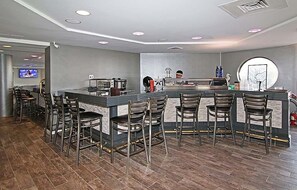 A modern bar area with a counter and multiple stools, a television screen in the background, and a large mirror on the wall.