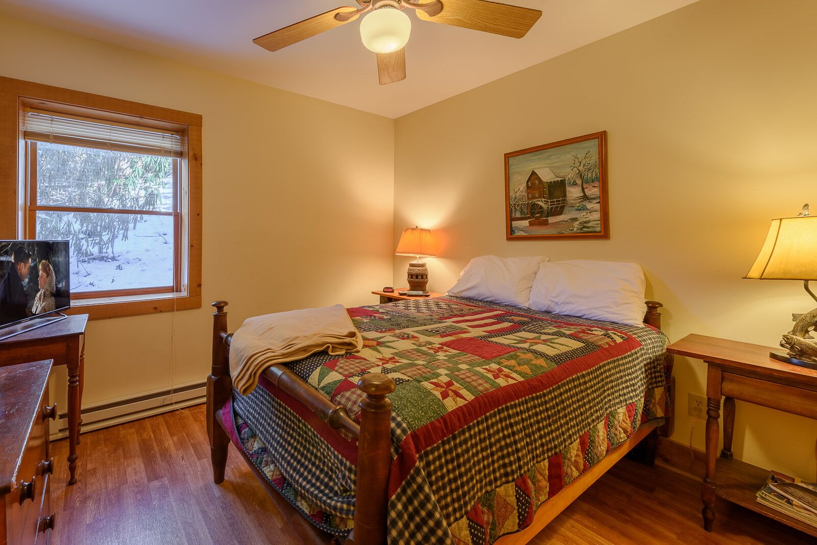 A bedroom with a wooden bed, a quilt, a painting, a lamp, and a TV on a wooden table.