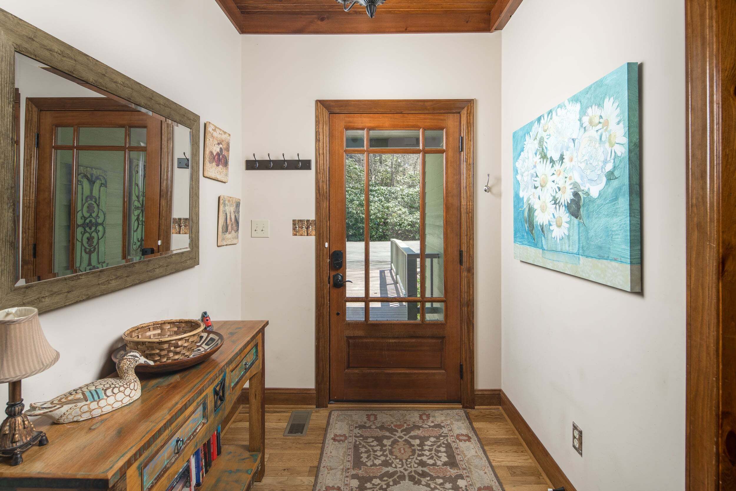 Une porte en bois avec des panneaux de verre, un miroir au-dessus, un tableau représentant des fleurs, une console en bois avec une lampe et un tapis à motifs.