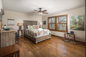 A bedroom with a wooden bed, a ceiling fan, a window with a forest view, a desk with a lamp, and a wall-mounted artwork.