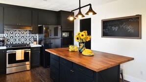 Una cocina moderna con armarios oscuros, una isla de madera y una pared de pizarra con un mensaje de bienvenida.