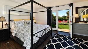 Una cama con dosel con una colcha floral, una mesita de noche con lámpara, un aparador con espejo y vista a un jardín a través de un ventanal.