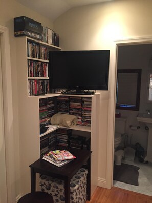 A room with a TV on a stand, a bookshelf filled with DVDs, a small table with magazines, and a cat sleeping on the shelf.