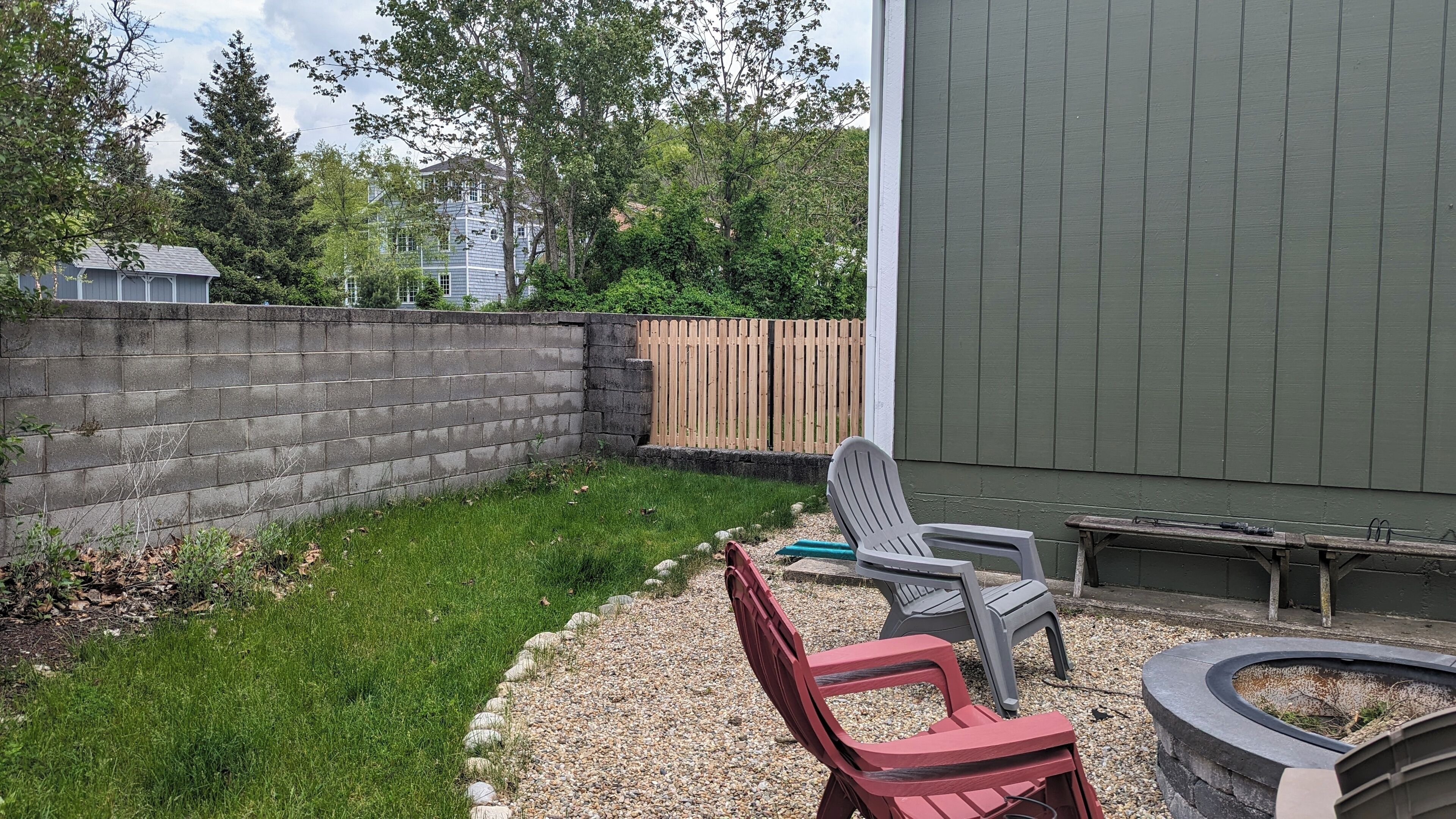 Un jardin avec un mur en béton, une clôture en bois et une zone herbeuse. On y trouve deux chaises et un petit foyer.
