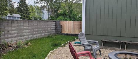 Un jardin avec un mur en béton, une clôture en bois et une zone herbeuse. On y trouve deux chaises et un petit foyer.
