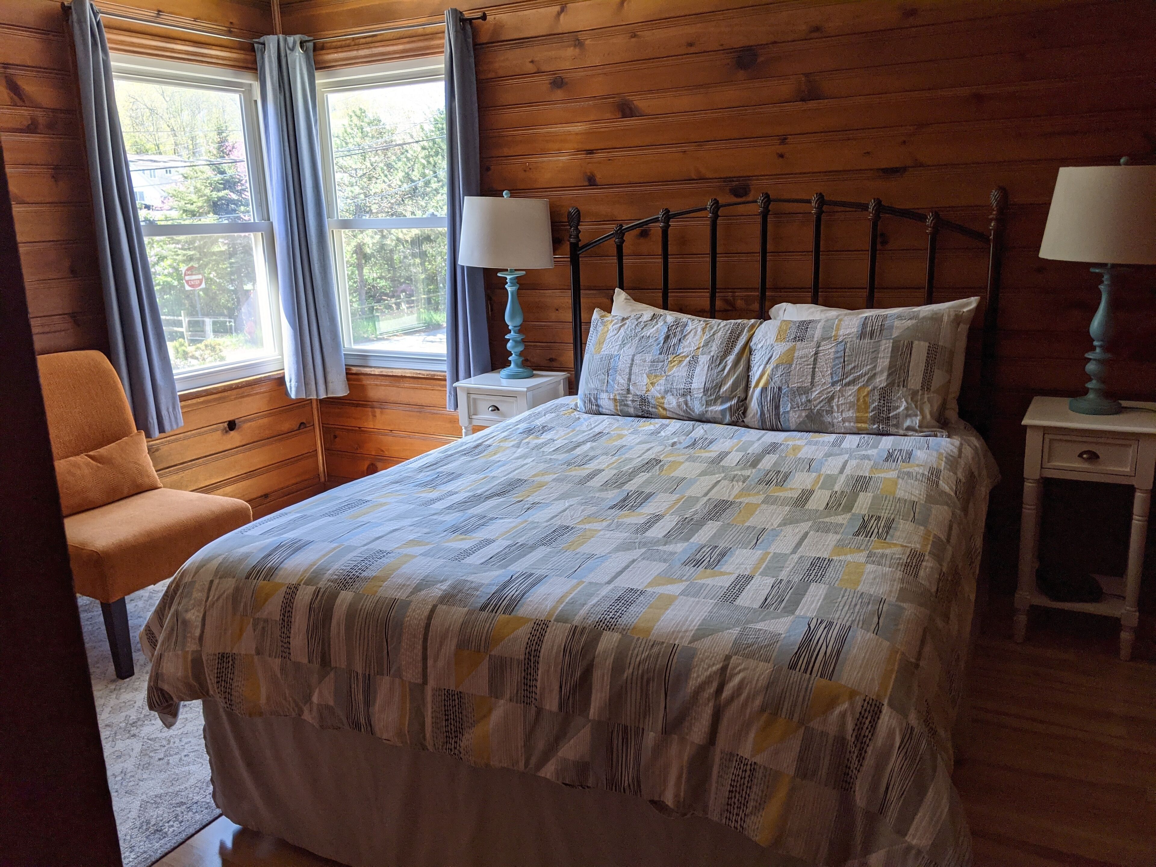 A bedroom with a wooden headboard, a bed with patterned bedding, two bedside tables with lamps, a chair, and a window with curtains.