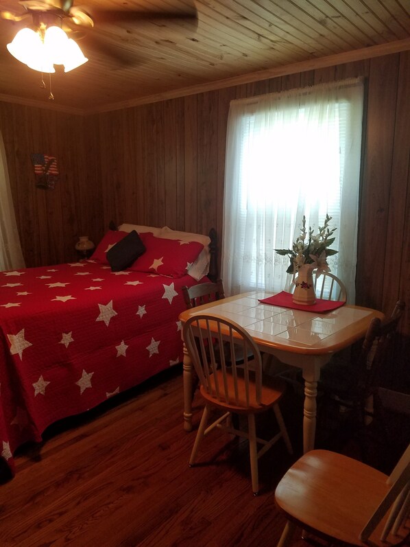 Una habitación con techo de madera, una cama con manta roja, una mesa con sillas y una ventana con cortinas.
