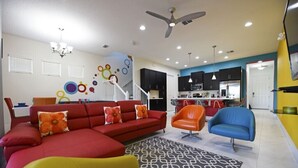 A modern living room with a red sectional sofa, orange and blue chairs, a patterned rug, and a kitchen in the background. - Festive Family Fun! Resort Close 2 Disney! Private Pool! 422 (Davenport)