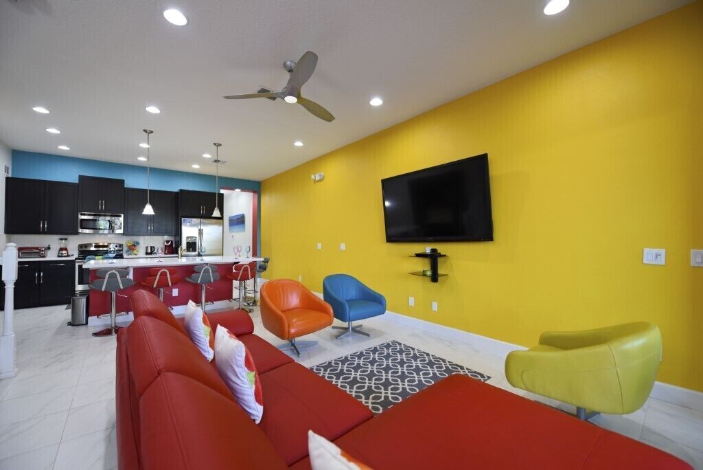 Una sala de estar moderna con sofás rojos, una pared de acento amarilla, un televisor de pantalla plana y una cocina al fondo.