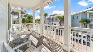 A covered porch with white railings and Adirondack chairs, overlooking a street with houses and palm trees.
