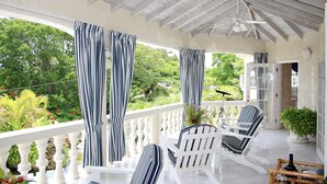 Un espace extérieur aménagé avec des sièges, protégé par un toit, comprenant des chaises blanches et des rideaux à rayures, et offrant une vue sur un jardin luxuriant.