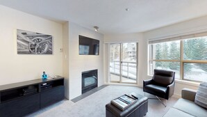 A modern living room with a fireplace, a black leather chair, a flat-screen TV, and a large window with a view of a snowy landscape. - Aspens #335 Prime Ski-in Ski-out Location! Pool, Hot tubs, BBQ, sleeps 4 (Whistler)