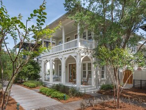 Une maison à deux étages dotée d’un porche abrité, d’une terrasse en bois et d’un jardin où poussent diverses plantes et arbres.