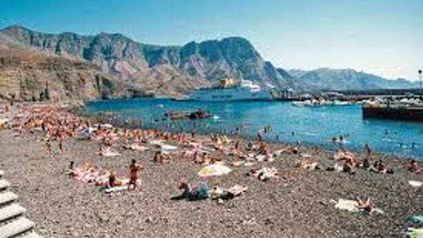 Ein Strand mit badenden und sonnenbächelnden Personen, felsigem Gelände und Bergen im Hintergrund.