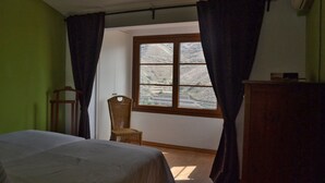 Ein Schlafzimmer mit einem Bett, einem Sessel, einem Fenster mit Vorhängen und Blick auf eine gebirgige Landschaft.
