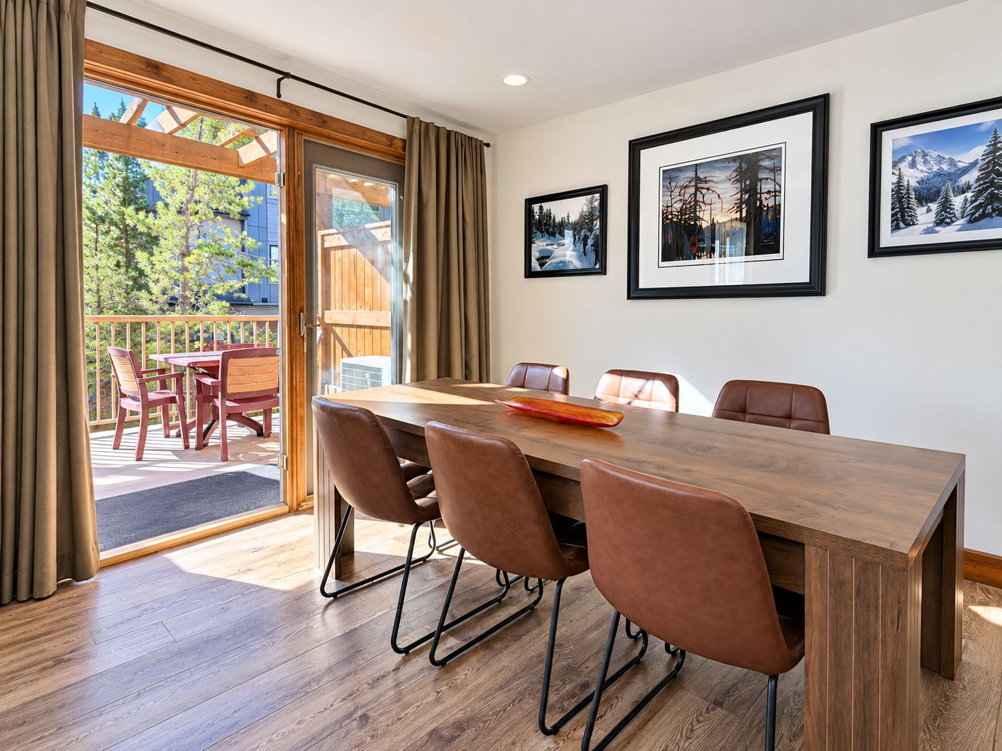 A dining area with a wooden table and chairs, a view of the outdoors through a glass door, and framed pictures on the wall.
