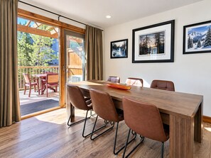 Un comedor con mesa y sillas de madera, vistas al exterior a través de un ventanal y cuadros en la pared.