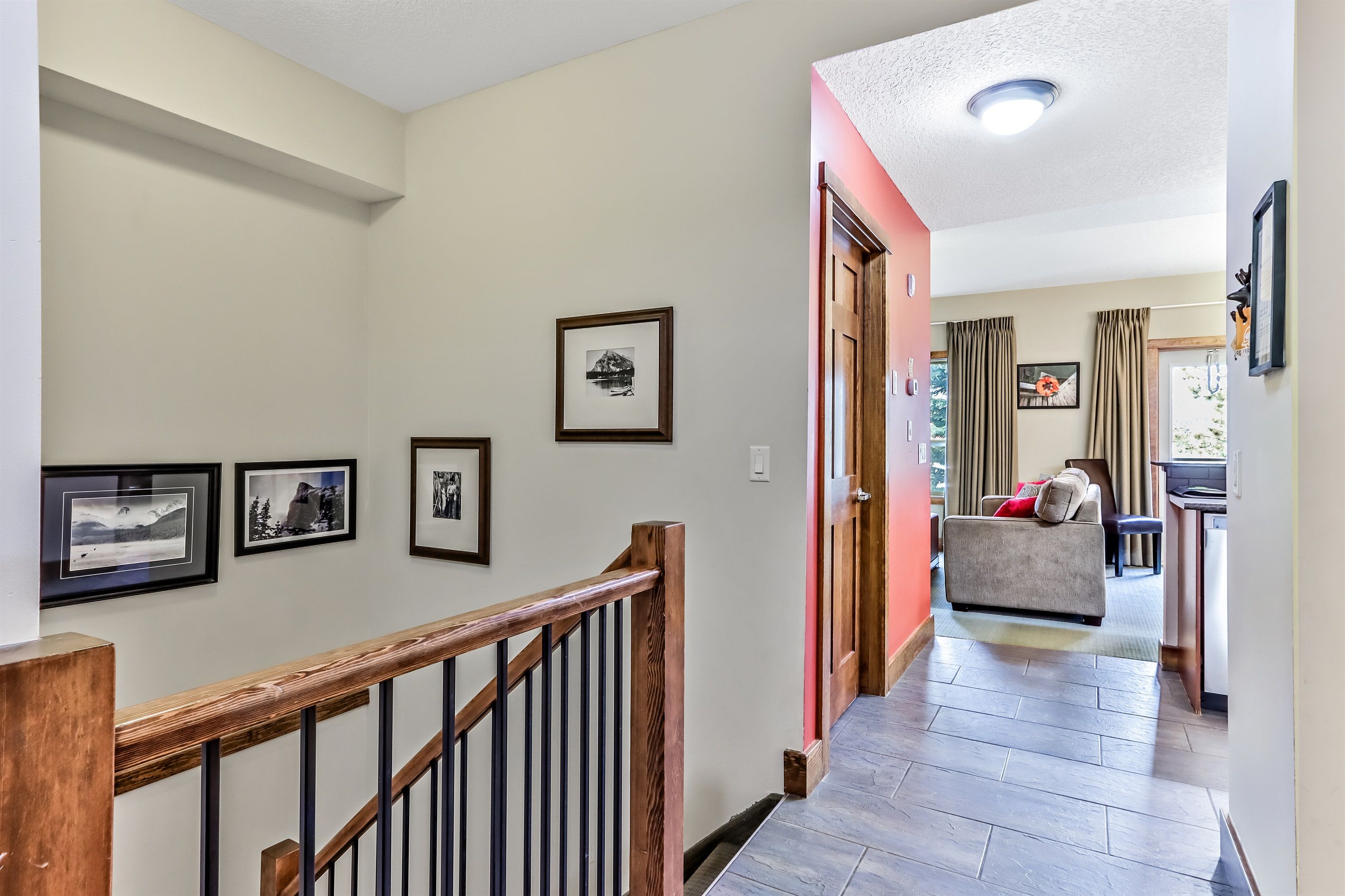 A hallway with framed pictures on the wall, a wooden railing, and a door leading to a living area with a sofa and curtains.