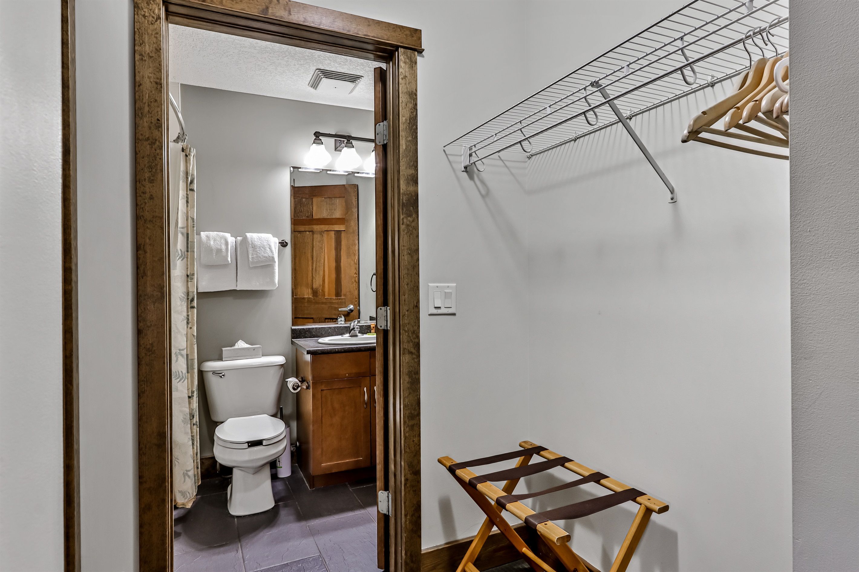 Ein Badezimmer mit Toilette, Waschbecken und Holzausbau. Ein Wäscheständer mit Kleidung und ein hölzerner Hocker sind ebenfalls vorhanden.