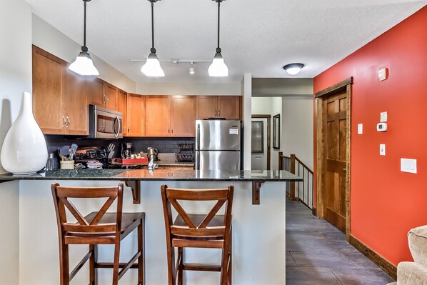A kitchen with wooden cabinets, stainless steel appliances, and a central island with two wooden chairs. - Spacious, Pet Friendly, Townhouse – Hot Tub | Perfect for Families! (Canmore)