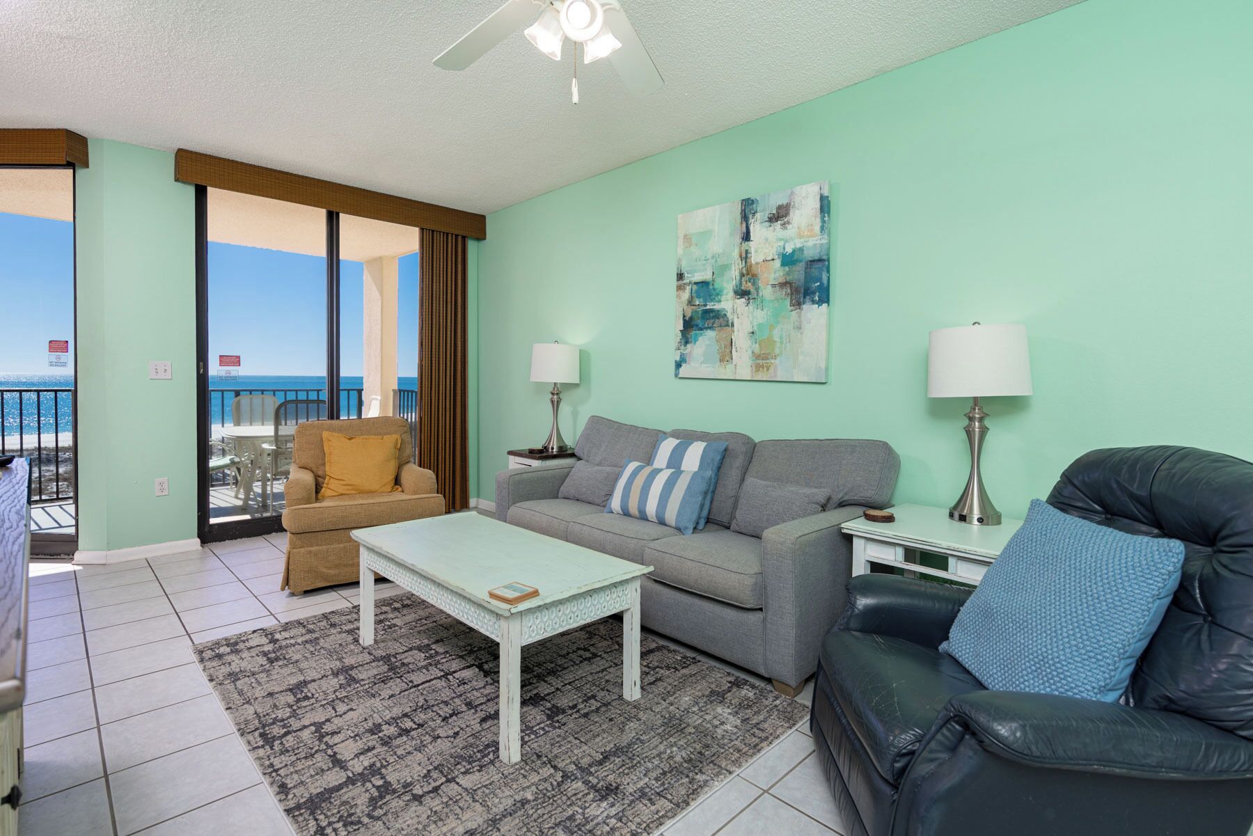 A living room with a sofa, armchair, and coffee table, a painting on the wall, and a view of the ocean through a sliding door.