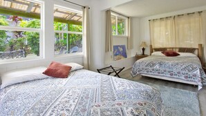 Una habitación con dos camas, cada una con ropa de cama estampada y un cojín rojo. Hay ventanas con cortinas, una silla y un cuadro en la pared.